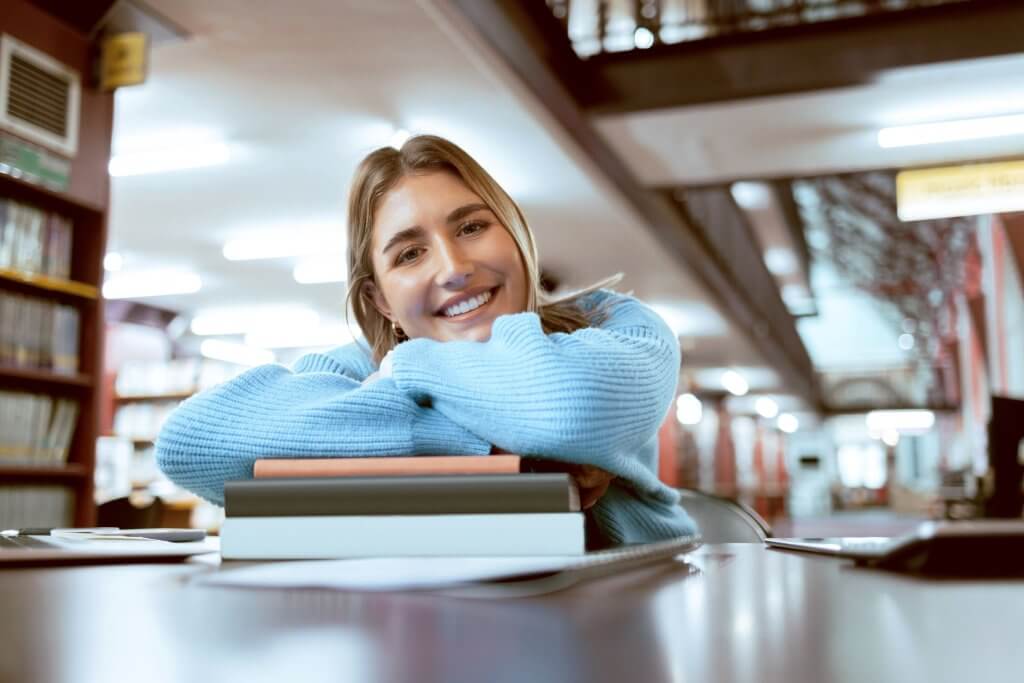Junge duale Studentin lächelt beim Lernen in der Bibliothek in die Kamera.