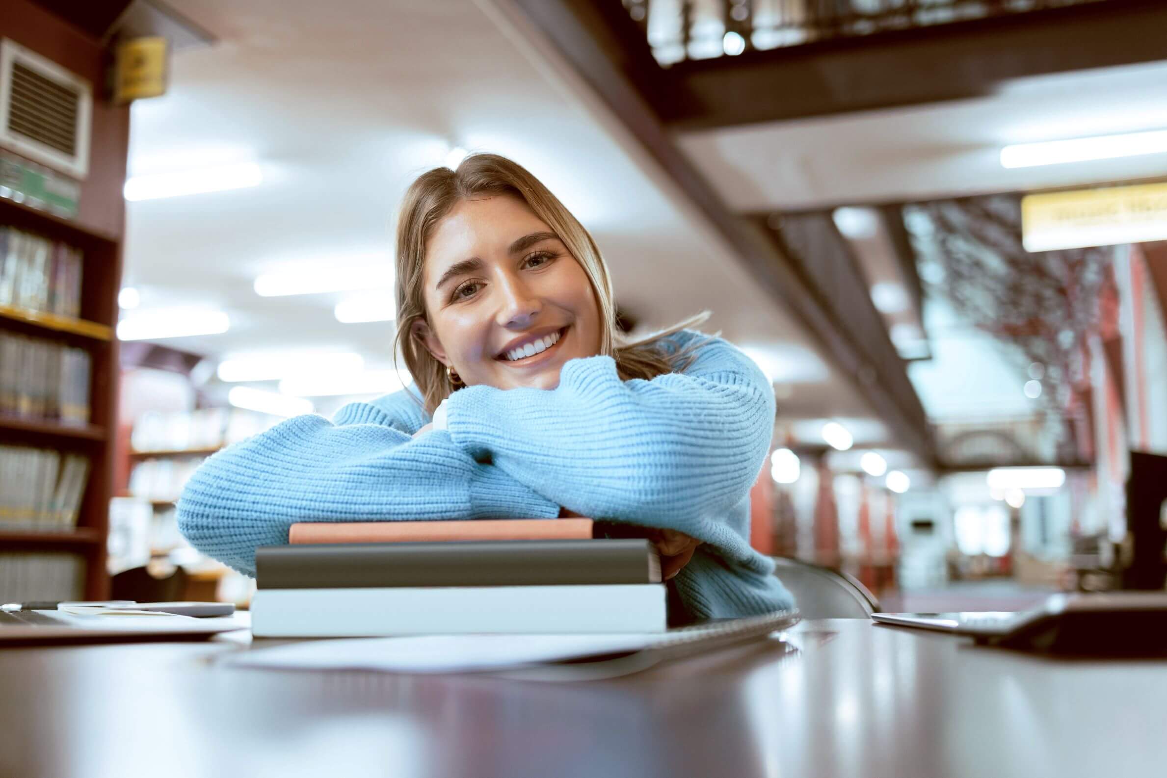 Junge duale Studentin lächelt beim Lernen in der Bibliothek in die Kamera.